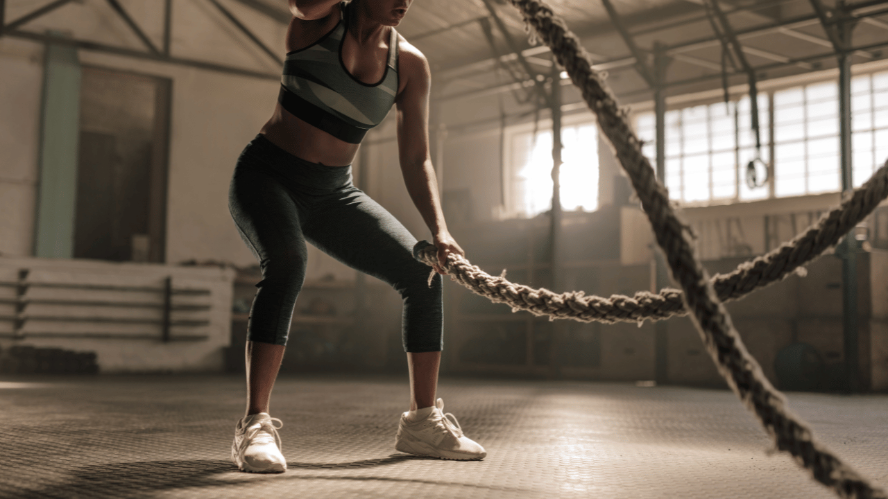 Female athlete performing high-intensity battle rope training in a gym environment during repeated efforts and heat-generating conditioning work.