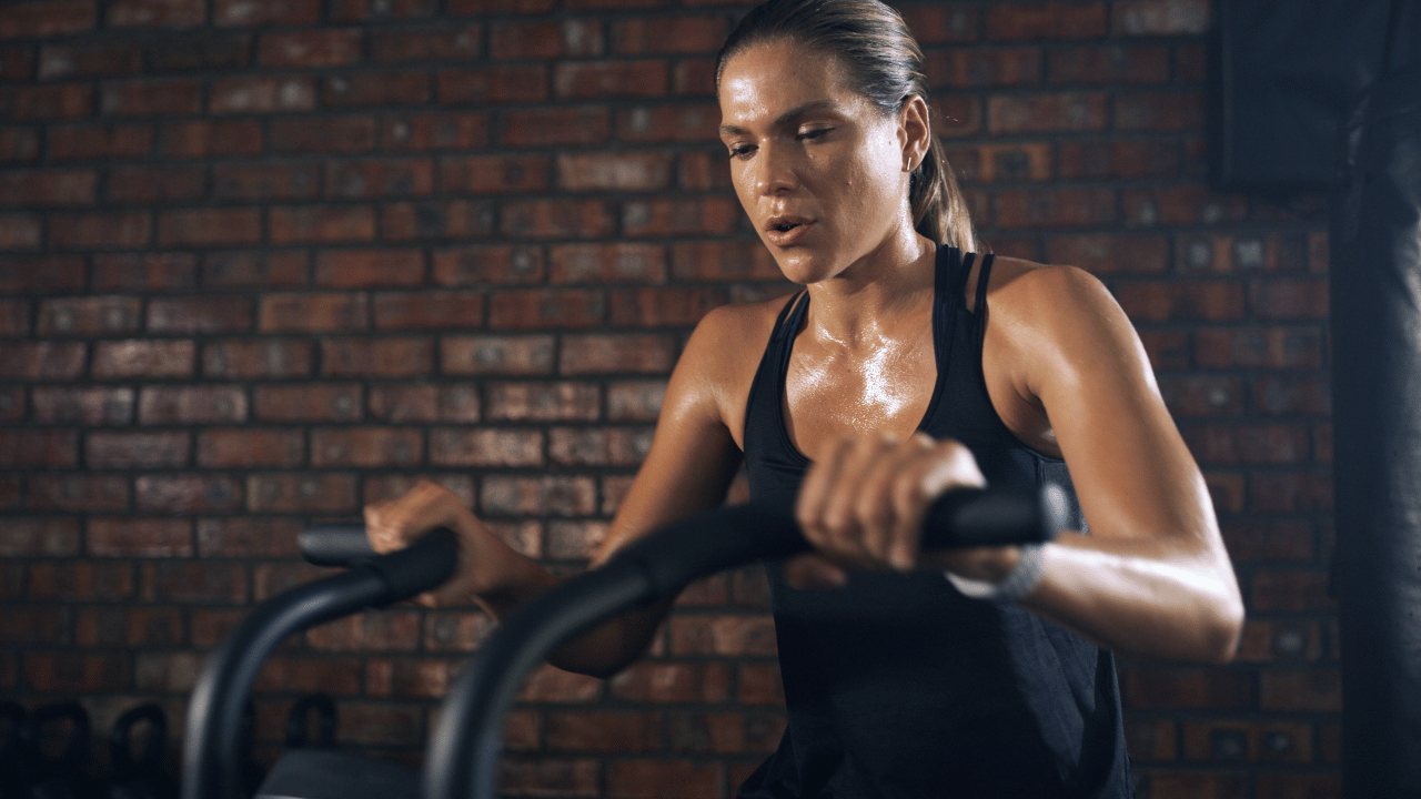 Female athlete covered in sweat during an intense assault bike workout, illustrating heat accumulation and repeated high-output training.